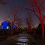 Een griezelige, schemerige scène toont een pad dat naar een oud huis leidt. De ramen van het huis gloeien met blauw licht en kale bomen aan beide kanten werpen rode tinten. Een gloeiende witte bol zweeft bij het huis, wat een surrealistische sfeer creëert.