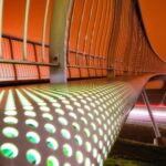 Een futuristische, verlichte brug met geperforeerde, gloeiende panelen en een gebogen ontwerp strekt zich uit over een donker landschap. De lucht gloeit oranje, wat een dramatische achtergrond creëert. Vangrails staan langs de brug en lichtstrepen van passerende voertuigen zijn zichtbaar beneden.