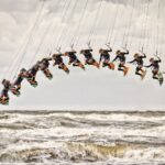 Een sequentie van de sprong van een kitesurfer, weergegeven in meerdere frames, die een vloeiende boog tegen een bewolkte lucht creëren. De golven van de oceaan zijn ruw en de kleurrijke uitrusting van de kitesurfer contrasteert met de gedempte tonen van de zee en de lucht.