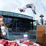 Een jonge vrouw zit aan een tafeltje buiten bij McDonald's, nippend aan een drankje en met frietjes in haar hand. Achter haar voert iemand een indrukwekkende salto uit in de lucht. De scène bevat McDonald's-voedselverpakkingen en een modern exterieur met het McDonald's-logo boven de ingang.