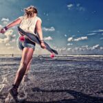 Een persoon in een wit shirt en een korte spijkerbroek rent door ondiep water op een strand met een skateboard in zijn hand. De oceaan en een gedeeltelijk bewolkte lucht zijn op de achtergrond te zien.