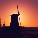 Silhouetten van windmolens steken hoog af tegen een levendige oranje en paarse zonsonderganghemel. De scène legt een kalm landelijk landschap vast met de contouren van bomen aan de horizon, wat een rustige, pittoreske avond oproept.