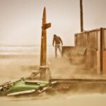 Een eenzame persoon staat op een houten platform in een winderige, zanderige strandomgeving. Een gedeeltelijk begraven groene jetski en een houten bord met een plank staan op de voorgrond. De scène is wazig en baadt in gedempte, aardse tinten.