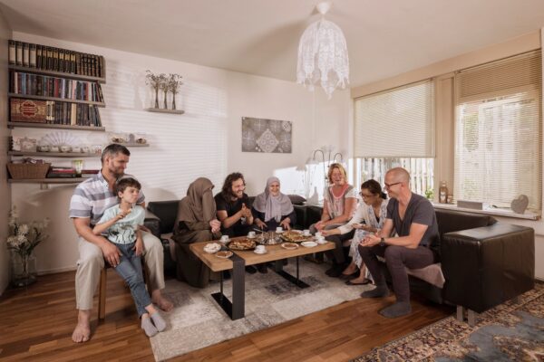 Een groep van acht personen, waaronder volwassenen en kinderen, zitten rond een salontafel in een gezellige woonkamer. De tafel staat vol met verschillende gerechten. De kamer heeft een warme ambiance met boekenplanken, kunst en natuurlijk licht dat door de jaloezieën naar binnen stroomt.