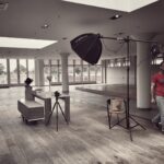 A spacious, empty room with wooden floors lit by natural light from large windows. A person sits at a desk with a camera on a tripod and studio light stand aimed at them. Another individual wearing a red shirt walks across the room.