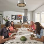 Vier mensen zitten rond een eettafel in een moderne keuken, delen een maaltijd en raken in gesprek. De keuken heeft een kookeiland versierd met bloemen, posters aan de muur en grote ramen die natuurlijk licht de ruimte laten vullen.