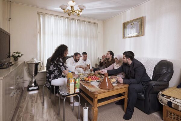 Een groep van zes personen zit rond een lage houten tafel in een gezellige woonkamer en deelt een maaltijd. Verschillende gerechten, drankjes en een tajinepot staan op tafel. De kamer is helder verlicht met een kroonluchter erboven en er staat een grote trofee op een nabijgelegen kast.