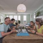 Een gezin van vier zit rond een houten eettafel in een moderne, goed verlichte kamer. Ze eten samen, lachend en pratend. De kamer heeft lichtgroene muren, ingelijste foto's, kamerplanten en eigentijds meubilair.