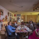 Een diverse groep mensen zit rond een lange eettafel en deelt een maaltijd in een warm ingerichte woonkamer. De tafel staat vol met verschillende gerechten. Kerstversieringen, waaronder slingers en kerstversiering, geven de kamer een feestelijke sfeer. Iedereen lijkt betrokken en gelukkig.