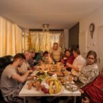 Een groep mensen zit rond een rechthoekige tafel vol met verschillende gerechten, fruit en drankjes. Het lijkt erop dat ze samenkomen voor een maaltijd in een woonkamer. De kamer heeft een witte muur, een klok, een bank en een gordijn met gouden tinten.