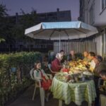 Een gezin is verzameld rond een tafel buiten, en eet onder een grote parasol. De tafel is bedekt met een groen gestreept kleed en verschillende gerechten. De scène speelt zich af in een achtertuin bij zonsondergang, met een gebouw en groen op de achtergrond.