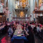 Een grote groep mensen zit aan lange tafels bedekt met blauwe tafellakens in een grote, sierlijke hal met hoge, gewelfde plafonds en kroonluchters. De kamer is helder verlicht en de sfeer lijkt feestelijk en levendig.