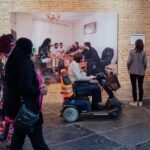 Een man op een scootmobiel met een oranje veiligheidsvest stopt om een grote foto te bekijken die op een bakstenen muur hangt. Twee vrouwen, een in een bloemenjurk en een ander in een lichtgekleurd jasje, bekijken het kunstwerk ook. De scène speelt zich af in wat een galerie lijkt te zijn.