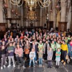 Een grote groep mensen van verschillende leeftijden, etniciteiten en vaardigheden verzamelt zich in een grote hal met sierlijke architectuur en kroonluchters. Sommigen lachen en poseren voor de foto, terwijl anderen in verschillende richtingen kijken. De setting ademt een levendige en diverse sfeer uit.