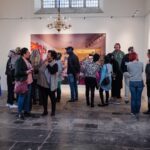 Een groep mensen met koptelefoons staat en communiceert in een kunstgalerie. De ruimte heeft hoge plafonds, grote ramen en een kroonluchter. Een grote ingelijste foto hangt op de muur op de achtergrond. De aanwezigen lijken betrokken en aandachtig.