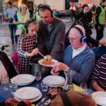 Mensen van verschillende leeftijden zitten rond een eettafel. Een man in pak helpt een jong meisje met het serveren van eten. Iedereen draagt een koptelefoon. De tafel is gedekt met borden, glazen en een waterkan. De achtergrond toont meer mensen in een gemeenschappelijke setting.