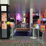 Een kleurrijke arcade-ingang met verschillende spelmachines zichtbaar binnenin. Er zijn ballonnen en posters met tekst bij de ingang, en een bord op de vloer met de tekst "PRIZEN CIRCUITUS." Felle lichten verlichten de ruimte.