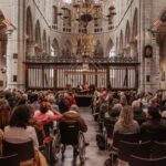 Een divers publiek zit in rijen in een grote, historische kerk met hoge gewelfde plafonds en luistert naar een luidsprekerpaneel aan de voorkant. Kroonluchters hangen aan het plafond en de omgeving wordt verlicht met warme verlichting. Een rolstoelgebruiker is zichtbaar tussen de aanwezigen.