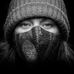 Zwart-wit close-up portret van een persoon die een gebreide muts en een gepatterned gezichtsmasker draagt. De achtergrond is volledig zwart, waardoor de aandacht wordt gevestigd op hun expressieve ogen en de textuur van zowel de muts als het masker.