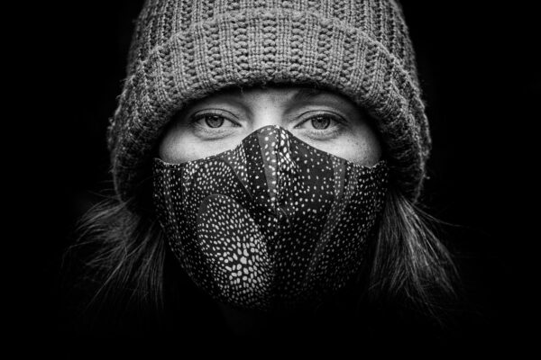 Zwart-wit close-up portret van een persoon die een gebreide muts en een gepatterned gezichtsmasker draagt. De achtergrond is volledig zwart, waardoor de aandacht wordt gevestigd op hun expressieve ogen en de textuur van zowel de muts als het masker.