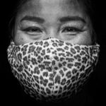 Een persoon lacht met zijn ogen terwijl hij een luipaardprint gezichtsmasker draagt. De achtergrond is zwart en de foto is in zwart-wit, waardoor de ogen en het masker van de persoon worden benadrukt.