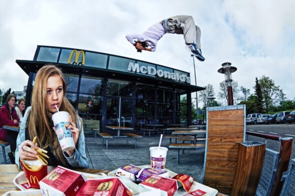 Een jong meisje zit aan een tafeltje buiten bij een McDonald's restaurant, nippend aan een drankje en frietjes etend. Op de achtergrond is een persoon in de lucht te zien die een salto uitvoert. Mensen zijn zichtbaar in het restaurant en in de rij buiten.