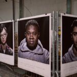 Een serie portretten tentoongesteld op een hek in een tentoonstellingsomgeving. De afbeeldingen tonen diverse personen; elk wordt vergezeld door tekst onder hun respectievelijke foto's. De achtergrond bevat industriële elementen zoals metalen staven en betonnen muren.