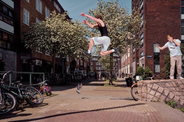 Een persoon in een wit tanktopje en een zwarte short is in de lucht en springt tussen twee verhoogde oppervlakken op een zonnige woonstraat. Een ander persoon staat op een van de oppervlakken, kijkt toe en gebaart. Fietsen staan in de buurt geparkeerd en bomen staan op de achtergrond.