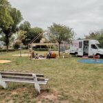 In een klein park staan een witte bank, twee ronde speelplekken met kleurrijke ontwerpen, een open houten constructie met zitplaatsen en een foodtruck met het opschrift 'Thuis Buik'. Onder de houten constructie zitten twee mensen, met huizen en bomen op de achtergrond.