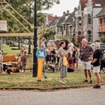 Een levendige straatbijeenkomst met mensen die kletsen, eten en drinken. Er zijn verschillende zitgelegenheden, waaronder fauteuils en een bank onder een houten pergola met een lamp eraan. Fietsen en geparkeerde auto's zijn zichtbaar aan de kant van de straat.