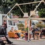 Een informeel zitgedeelte buiten is ingericht met een houten frameconstructie, banken en stoelen. Een groep mensen zit en staat, praat en ontspant. In de buurt is een vrachtwagen met tekst zichtbaar en er staan scooters geparkeerd. Bomen en gebouwen omringen de scène.