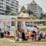 Een groep mensen verzamelt zich rond een tafel onder een houten frameconstructie met hangende lampenkappen, opgesteld naast een witte mobiele bus. De bus heeft kleurrijke afbeeldingen en staat geparkeerd op een grasveld, met hoge woongebouwen op de achtergrond.