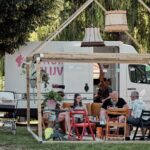 Een groep mensen is verzameld onder een houten pergola met hangende lampen, zittend op kleurrijke stoelen rond een tafel. Een witte bestelwagen met gedeeltelijk onleesbare tekst staat achter hen geparkeerd. Bomen en groen omringen de scène, wat suggereert dat het een ongedwongen, buitenomgeving is.