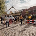 Mensen zijn verzameld op een geplaveide straat met stoelen onder een eenvoudige houten constructie. In de buurt is er een barricade met een bord, wat tafels met spullen, een geparkeerde bus en huizen. Het tafereel wordt verlicht door daglicht en versierd met kleine vlaggetjes.