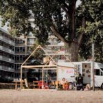 Een camperbusje geparkeerd naast een grote boom voor een modern appartementengebouw. Naast het busje staat een houten frameconstructie, gedeeltelijk bedekt met een luifel. Meerdere mensen zitten of spelen rond het frame en het busje, genietend van een zonnige dag.