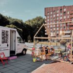 Een mobiele bus van een buurthuis staat geparkeerd naast een geïmproviseerd zitgedeelte buiten. Meerdere mensen zitten aan een houten tafel onder een frame dat lijkt op een huis. De achtergrond toont een bakstenen appartementengebouw met meerdere verdiepingen en een heldere, zonnige lucht.