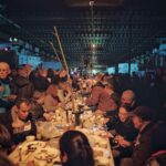 Een grote menigte mensen verzamelt zich aan een lange, overdekte feesttafel, genietend van eten en drinken. Lichtsnoeren en decoraties hangen aan het plafond. De sfeer is feestelijk en levendig, met mensen die praten en eten in een warm verlichte ruimte.