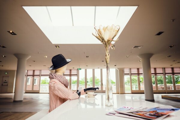 Een mannequin gekleed in een roze jas, sjaal en zwarte hoed zit aan een moderne, witte toonbank met een kop en schotel. Een hoge vaas met een grote bloem staat op de toonbank. Tijdschriften liggen verspreid op de voorgrond. Grote ramen en dakramen verlichten de ruimte.