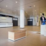 An empty retail store features a long checkout counter with "kassa" signs above. The room is brightly lit with minimalist decor, including a bench in the forefront and a mannequin dressed in blue near the center. A tree design decorates the wall behind the counter.