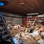 Een rommelige kamer is gevuld met verspreide dozen, papieren en boekenplanken. Verschillende menselijke figuren, gedeeltelijk bedekt met puin, zijn zichtbaar in de chaos. Een houten plafond en een ladder die tegen de linkermuur leunen, bieden extra context aan de wanorde van de scène.