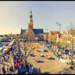 Een druk marktplein in een Europese stad met een prominente kerk of kathedraal op de achtergrond. Talrijke kraampjes staan langs het plein en een menigte mensen loopt rond, bezig met verschillende activiteiten. De afbeelding heeft een tilt-shift-effect, waardoor een miniatuur-achtig uiterlijk ontstaat.