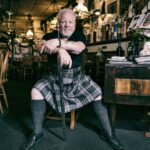 Een man met wit haar en een baard zit zelfverzekerd aan een tafel in een rustieke pub of restaurant. Hij draagt een traditionele tartan kilt en houdt een wandelstok vast. De gezellige setting bestaat uit houten meubels, verschillende decoraties en planken vol flessen en memorabilia.