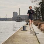 Een persoon jogt op een heldere dag langs een pad langs het water. Ze dragen zwarte sportkleding terwijl ze rennen op het betonnen pad, met water aan de ene kant en gebouwen op de achtergrond. Struiken en een boom zijn zichtbaar langs het pad.