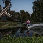 Een persoon met een zwemvest is aan het waterskiën op een smal water. De achtergrond toont een kleine traditionele windmolen, bomen en huizen. De beweging van de skiër creëert een spray van water, wat duidt op snelheid en vaardigheid. Het tafereel lijkt zich af te spelen in de schemering.