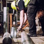 Een bouwvakker met een gele helm en veiligheidsvest gebruikt een slijpgereedschap om een metalen pijp door te snijden, waardoor er vonken vliegen. Verschillende metalen pijpen liggen verspreid op de grond om hen heen en de scène speelt zich buiten af op een zonnige dag.