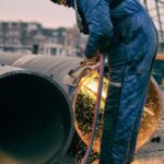 Een bouwvakker in een blauwe overall, helm en handschoenen gebruikt een snijbrander om door een grote metalen pijp te snijden. Er vliegen vonken van de brander terwijl de arbeider, staand buiten op een betonnen oppervlak, zich concentreert op de taak. Stedelijke structuren zijn zichtbaar op de achtergrond.