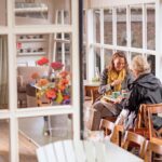 Twee vrouwen zitten aan een houten tafel in een gezellig café met grote ramen, delen een maaltijd en lachen. Het café is versierd met levendige bloemen en zachte stoelen. De sfeer lijkt warm en uitnodigend, met natuurlijk licht dat door de ramen stroomt.