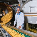 Een arbeider in beschermende kleding en haarnetje staat naast industriële machines die spiraalvormige pasta produceren. Hij glimlacht terwijl de pasta van een grote, cilindrische machine op een transportband stroomt. De setting lijkt een voedselproductiefaciliteit te zijn.