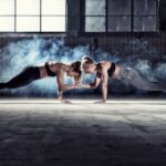 Twee vrouwen zijn bezig met een plankpositie terwijl ze op één hand balanceren en hun vrije handen in elkaar vouwen. Ze dragen sportkleding in een slecht verlichte sportschool met een groot raam en rook op de achtergrond, wat een dramatische sfeer creëert.