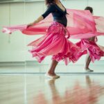 Een vrouw danst sierlijk met een roze, vloeiende rok in een dansstudio. Ze glimlacht, houdt de rok naar buiten, waardoor er een werveling van stof om haar heen ontstaat. Haar weerspiegeling is zichtbaar in de spiegelwand achter haar, en de houten vloer voegt warmte toe aan de scène.